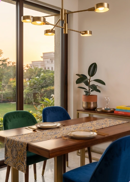 A vibrant, modern dining room setup featuring a rich wood table with an ornate runner, surrounded by jewel-toned blue and green velvet chairs, representing Craftnika's premium furniture trusted across India and global export markets.